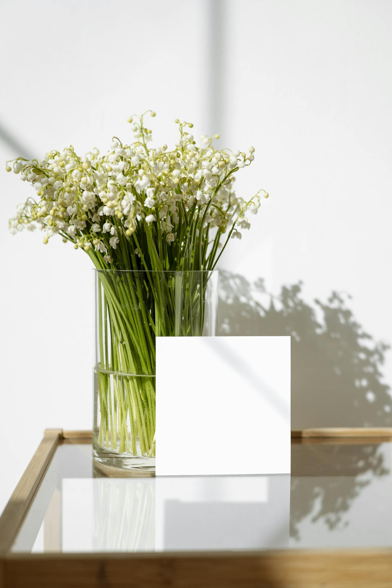 A minimalist arrangement of white flowers in a glass vase alongside a blank card on a glass table.